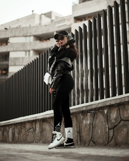 A person wearing urban streetwear poses confidently against a modern architectural background. The outfit includes a black cap, oversized sunglasses, a black leather jacket, and black leggings with white socks and sneakers, along with a white handbag. The setting features a tall, dark fence and a concrete structure in the background, creating a stylish, urban vibe.