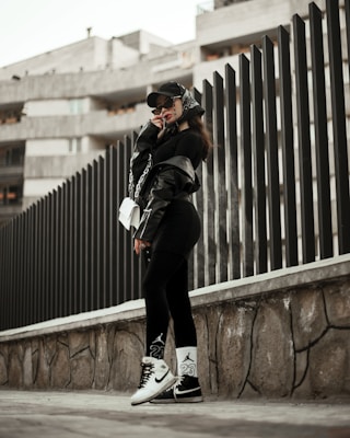 A person wearing urban streetwear poses confidently against a modern architectural background. The outfit includes a black cap, oversized sunglasses, a black leather jacket, and black leggings with white socks and sneakers, along with a white handbag. The setting features a tall, dark fence and a concrete structure in the background, creating a stylish, urban vibe.