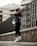 A person wearing urban streetwear poses confidently against a modern architectural background. The outfit includes a black cap, oversized sunglasses, a black leather jacket, and black leggings with white socks and sneakers, along with a white handbag. The setting features a tall, dark fence and a concrete structure in the background, creating a stylish, urban vibe.