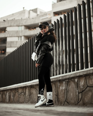 A person wearing urban streetwear poses confidently against a modern architectural background. The outfit includes a black cap, oversized sunglasses, a black leather jacket, and black leggings with white socks and sneakers, along with a white handbag. The setting features a tall, dark fence and a concrete structure in the background, creating a stylish, urban vibe.