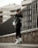 A person wearing urban streetwear poses confidently against a modern architectural background. The outfit includes a black cap, oversized sunglasses, a black leather jacket, and black leggings with white socks and sneakers, along with a white handbag. The setting features a tall, dark fence and a concrete structure in the background, creating a stylish, urban vibe.