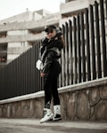 A person wearing urban streetwear poses confidently against a modern architectural background. The outfit includes a black cap, oversized sunglasses, a black leather jacket, and black leggings with white socks and sneakers, along with a white handbag. The setting features a tall, dark fence and a concrete structure in the background, creating a stylish, urban vibe.