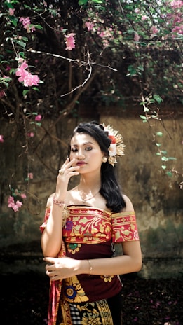 A woman stands elegantly amidst a backdrop of vibrant foliage, wearing traditional attire with intricate red and gold patterns. Her hair is adorned with decorative flowers, complementing the floral theme. The background features a rustic wall and blooming pink flowers, adding to the natural aesthetic.