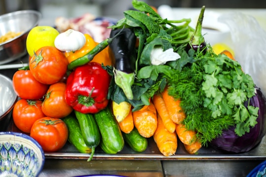 A vibrant assortment of fresh vegetables including tomatoes, cucumbers, carrots, red bell peppers, eggplant, garlic, and leafy greens is arranged on a tray. The produce appears fresh and colorful, with a variety of textures and shapes. The background includes other kitchen items slightly out of focus.