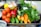 A vibrant assortment of fresh vegetables including tomatoes, cucumbers, carrots, red bell peppers, eggplant, garlic, and leafy greens is arranged on a tray. The produce appears fresh and colorful, with a variety of textures and shapes. The background includes other kitchen items slightly out of focus.