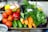 A vibrant assortment of fresh vegetables including tomatoes, cucumbers, carrots, red bell peppers, eggplant, garlic, and leafy greens is arranged on a tray. The produce appears fresh and colorful, with a variety of textures and shapes. The background includes other kitchen items slightly out of focus.