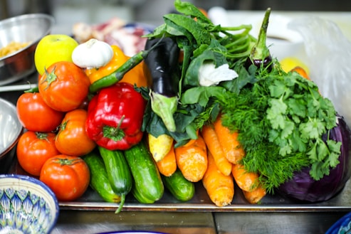 A vibrant assortment of fresh vegetables including tomatoes, cucumbers, carrots, red bell peppers, eggplant, garlic, and leafy greens is arranged on a tray. The produce appears fresh and colorful, with a variety of textures and shapes. The background includes other kitchen items slightly out of focus.