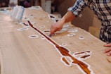 A person is working on a wooden table with artistic inlays, using a tool to shape or fill a resin river design. The surface is predominantly light-colored wood, and there are various white outlines possibly marking where resin will be placed.