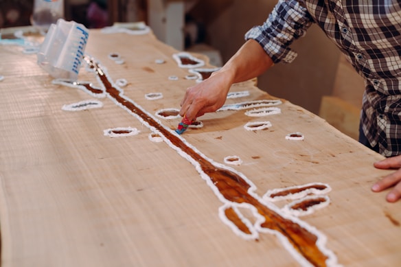 A person is working on a wooden table with artistic inlays, using a tool to shape or fill a resin river design. The surface is predominantly light-colored wood, and there are various white outlines possibly marking where resin will be placed.