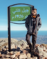 A person wearing a jacket and sunglasses stands on rocky terrain next to a green sign with Arabic text. The background reveals a vast landscape with mountains and a blue sky, leading to a distant ocean view.