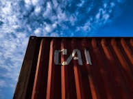 A red shipping container with the letters 'CAI' painted on its side is positioned against a backdrop of a blue sky filled with scattered white clouds. The image captures a perspective looking upwards at the container, highlighting the play of shadow and light across its surface.