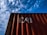 A red shipping container with the letters 'CAI' painted on its side is positioned against a backdrop of a blue sky filled with scattered white clouds. The image captures a perspective looking upwards at the container, highlighting the play of shadow and light across its surface.