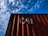A red shipping container with the letters 'CAI' painted on its side is positioned against a backdrop of a blue sky filled with scattered white clouds. The image captures a perspective looking upwards at the container, highlighting the play of shadow and light across its surface.