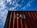 A red shipping container with the letters 'CAI' painted on its side is positioned against a backdrop of a blue sky filled with scattered white clouds. The image captures a perspective looking upwards at the container, highlighting the play of shadow and light across its surface.