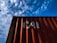 A red shipping container with the letters 'CAI' painted on its side is positioned against a backdrop of a blue sky filled with scattered white clouds. The image captures a perspective looking upwards at the container, highlighting the play of shadow and light across its surface.