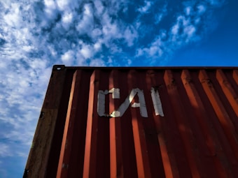 A red shipping container with the letters 'CAI' painted on its side is positioned against a backdrop of a blue sky filled with scattered white clouds. The image captures a perspective looking upwards at the container, highlighting the play of shadow and light across its surface.
