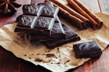 Several pieces of dark chocolate with embossed designs rest on a piece of brown paper. Cinnamon sticks are positioned nearby, and the whole arrangement sits on a wooden surface, creating a rustic presentation.
