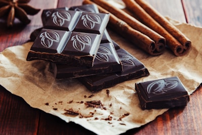 Several pieces of dark chocolate with embossed designs rest on a piece of brown paper. Cinnamon sticks are positioned nearby, and the whole arrangement sits on a wooden surface, creating a rustic presentation.