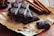 Several pieces of dark chocolate with embossed designs rest on a piece of brown paper. Cinnamon sticks are positioned nearby, and the whole arrangement sits on a wooden surface, creating a rustic presentation.