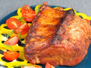 A perfectly roasted piece of meat served on a blue plate, garnished with yellow bell pepper rings and halved cherry tomatoes. The meat has a crispy, caramelized exterior with a rich brown color.