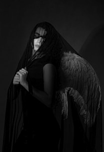 A person draped in a dark, translucent veil stands against a dark background. The figure appears contemplative, with eyes closed or downcast. The garment they wear is complemented by a large, detailed feathered wing on their back, suggesting a celestial or ethereal theme. The black and white composition adds depth and contrast to the image, highlighting the textures of the veil and feathers.