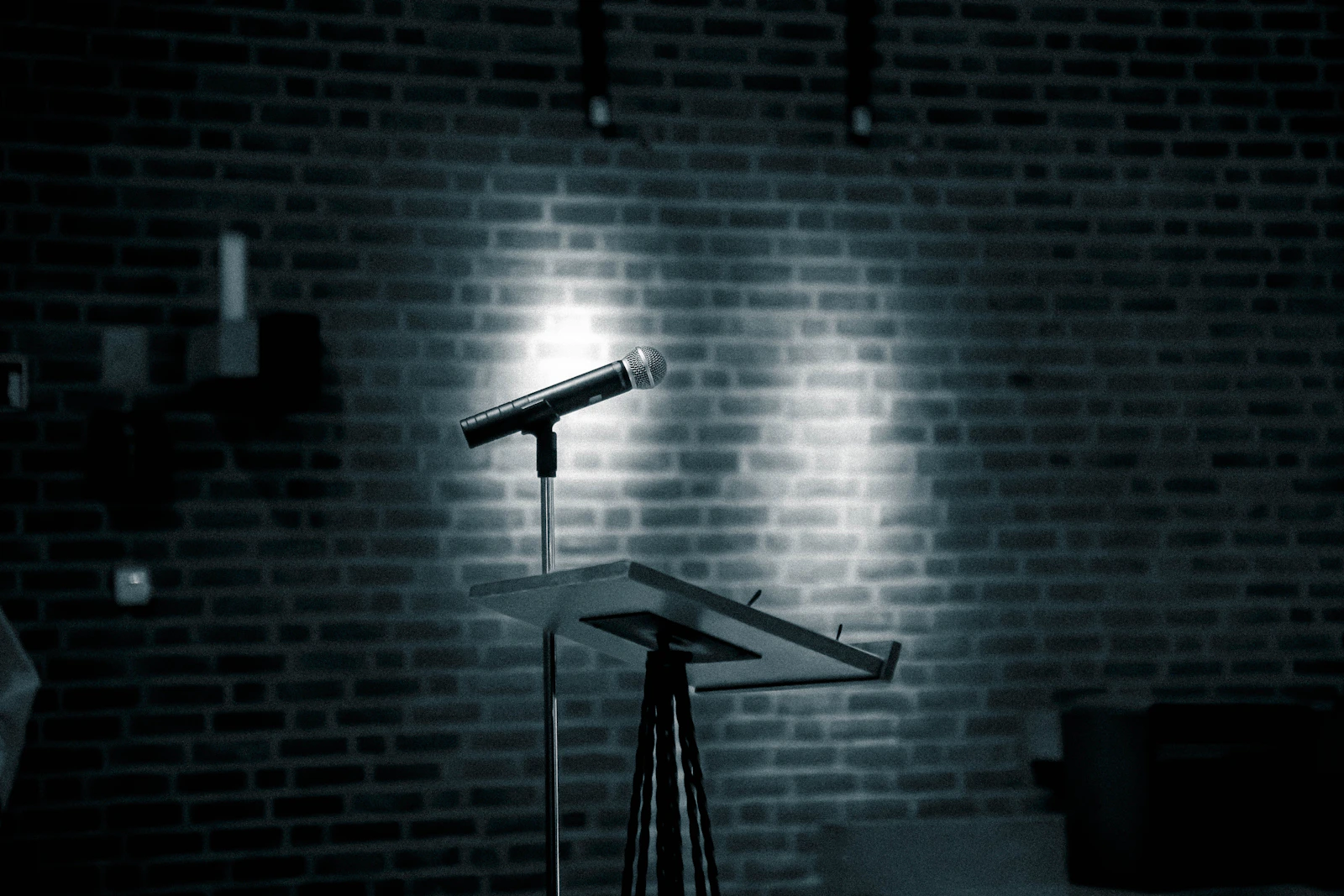 A microphone on a stand in front of a brick wall, a real stock photo of a stand-up comedy stage with no people in the frame, chosen for the Norm Macdonald documentary about a stand-up legend