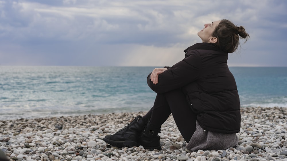 a woman sitting on a rocky beach looking up at the sky