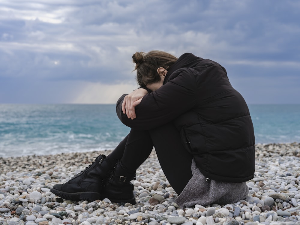 a woman sitting on a rocky beach with her head in her hands