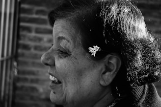 A grayscale image featuring a profile view of a woman smiling. Her hair is styled neatly with a small, delicate flower accessory. The background is composed of blurred, textured brickwork, providing a contrast to the focused, detailed profile.