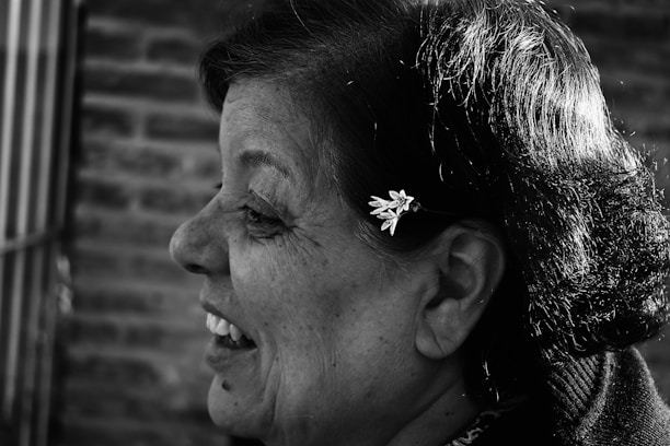 A grayscale image featuring a profile view of a woman smiling. Her hair is styled neatly with a small, delicate flower accessory. The background is composed of blurred, textured brickwork, providing a contrast to the focused, detailed profile.