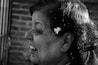 A grayscale image featuring a profile view of a woman smiling. Her hair is styled neatly with a small, delicate flower accessory. The background is composed of blurred, textured brickwork, providing a contrast to the focused, detailed profile.