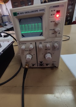 An oscilloscope displaying a green waveform on its screen, with various control knobs and switches, including power, intensity, focus, and calibration settings. Connected cables can be seen extending from the device, which is placed on a wooden table in a laboratory-like setting.
