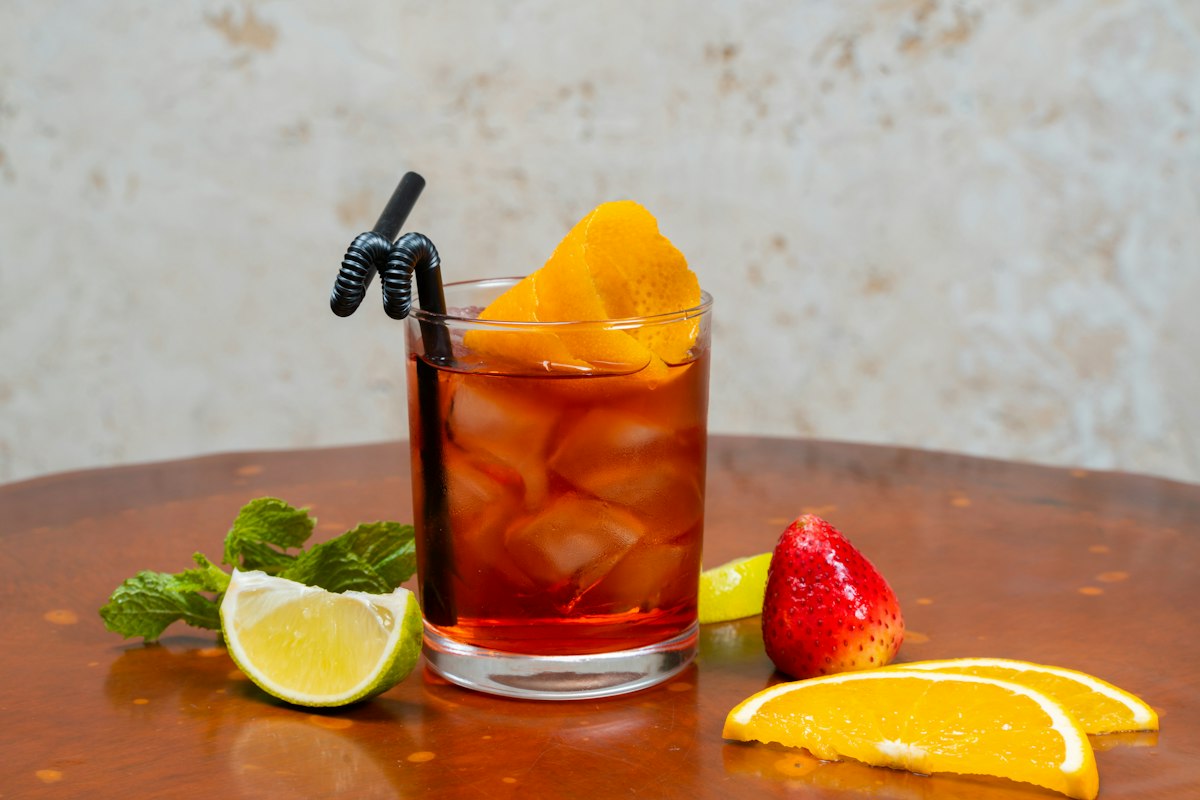 A glass of iced tea with ice cubes and an orange twist, surrounded by fresh mint, lime, strawberry, and orange slices on a wooden table