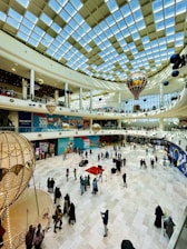 A large indoor shopping mall with a high glass ceiling allowing natural light. The floor is spacious, with groups of people walking or standing. Decorative structures resembling hot air balloons are suspended from the ceiling. Stores and advertisements are visible along the walls.