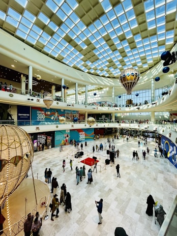 A large indoor shopping mall with a high glass ceiling allowing natural light. The floor is spacious, with groups of people walking or standing. Decorative structures resembling hot air balloons are suspended from the ceiling. Stores and advertisements are visible along the walls.