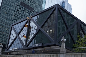A modern building with a geometric, crisscross steel frame surrounding large glass panels. The structure features an entrance sign for Fitness Formula Clubs. Nearby, there is a combination of sleek architectural elements typical of an urban environment, with tall skyscrapers in the background.