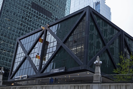 A modern building with a geometric, crisscross steel frame surrounding large glass panels. The structure features an entrance sign for Fitness Formula Clubs. Nearby, there is a combination of sleek architectural elements typical of an urban environment, with tall skyscrapers in the background.