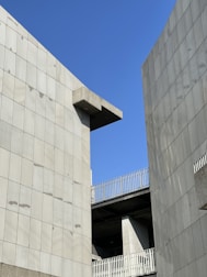 Concrete building structures with modern design elements set against a clear blue sky. The architecture features clean lines and a minimalist aesthetic, with the buildings forming a narrow gap through which the sky is visible.