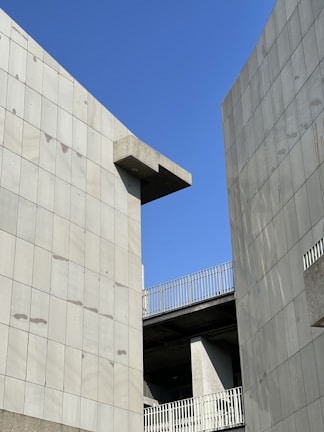 Concrete building structures with modern design elements set against a clear blue sky. The architecture features clean lines and a minimalist aesthetic, with the buildings forming a narrow gap through which the sky is visible.