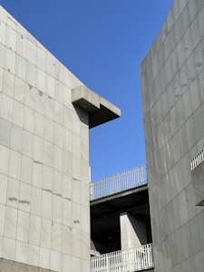 Concrete building structures with modern design elements set against a clear blue sky. The architecture features clean lines and a minimalist aesthetic, with the buildings forming a narrow gap through which the sky is visible.
