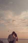 A couple stands close to each other under a partly cloudy sky, with their heads touching affectionately. There is a bird flying in the background over the ocean.