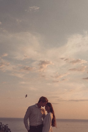A couple stands close to each other under a partly cloudy sky, with their heads touching affectionately. There is a bird flying in the background over the ocean.