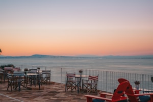 A scenic outdoor terrace with tables and chairs overlooks a vast body of water during a sunset. The sky transitions from soft pink to a gentle blue, offering a tranquil view. Empty tables and striped chairs are set on a tiled floor, with potted plants adding a touch of greenery.