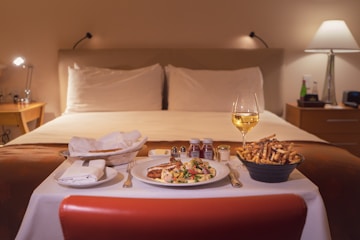 A hotel room setting with a neatly made bed in the background. In the foreground, a table set for a meal with a white tablecloth. There is a plate of pasta with vegetables, a bowl of fries, condiments, and a glass of white wine. The room is softly lit with a bedside lamp on a wooden nightstand.