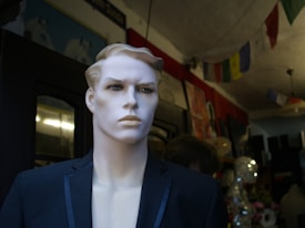 A mannequin with chiseled facial features wearing a dark suit is positioned in a dimly lit room. The background includes various hanging decorations and wall art, contributing to an eclectic atmosphere.