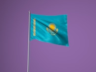 A flag with a sky blue background featuring a gold emblem of the sun with 32 rays above a soaring eagle. A vertical band with a national ornamental pattern is present on the left side of the flag. The flag is set against a solid purple background and is attached to a silver flagpole.