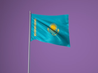 A flag with a sky blue background featuring a gold emblem of the sun with 32 rays above a soaring eagle. A vertical band with a national ornamental pattern is present on the left side of the flag. The flag is set against a solid purple background and is attached to a silver flagpole.