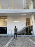 A person is standing on a stone pathway in front of a modern white building with gold accents, taking a photo of the building's facade. The facade features a set of stairs with gold railings leading to the upper floor and a neon sign that reads 'MYKITA.' There are large windows on the upper floor, and some plants are visible near the stairs.