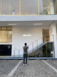 A person is standing on a stone pathway in front of a modern white building with gold accents, taking a photo of the building's facade. The facade features a set of stairs with gold railings leading to the upper floor and a neon sign that reads 'MYKITA.' There are large windows on the upper floor, and some plants are visible near the stairs.