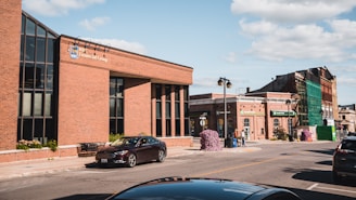 A street scene featuring a brick building with large windows on the left, identified as RBC Financial Group. There are several cars parked along the street, with one prominent black car nearby. The sky is clear with a few clouds. Further down the street, additional buildings are visible with one under renovation covered in green scaffolding. There are a couple of people walking on the sidewalk.